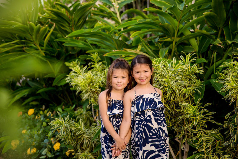 sisters holding hands in front of tropical plants and green foliage on the grounds of Ko Olina